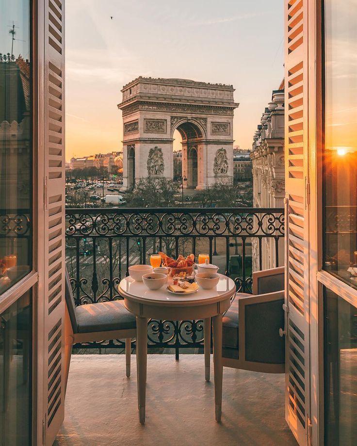 Cette image représente en premier plan une petite table pour deux personnes sur un balcon, et sur laquelle est servi un petit-déjeuner. En arrière plan on peut admirer la Place de l'Etoile avec l'Arc de triomphe 